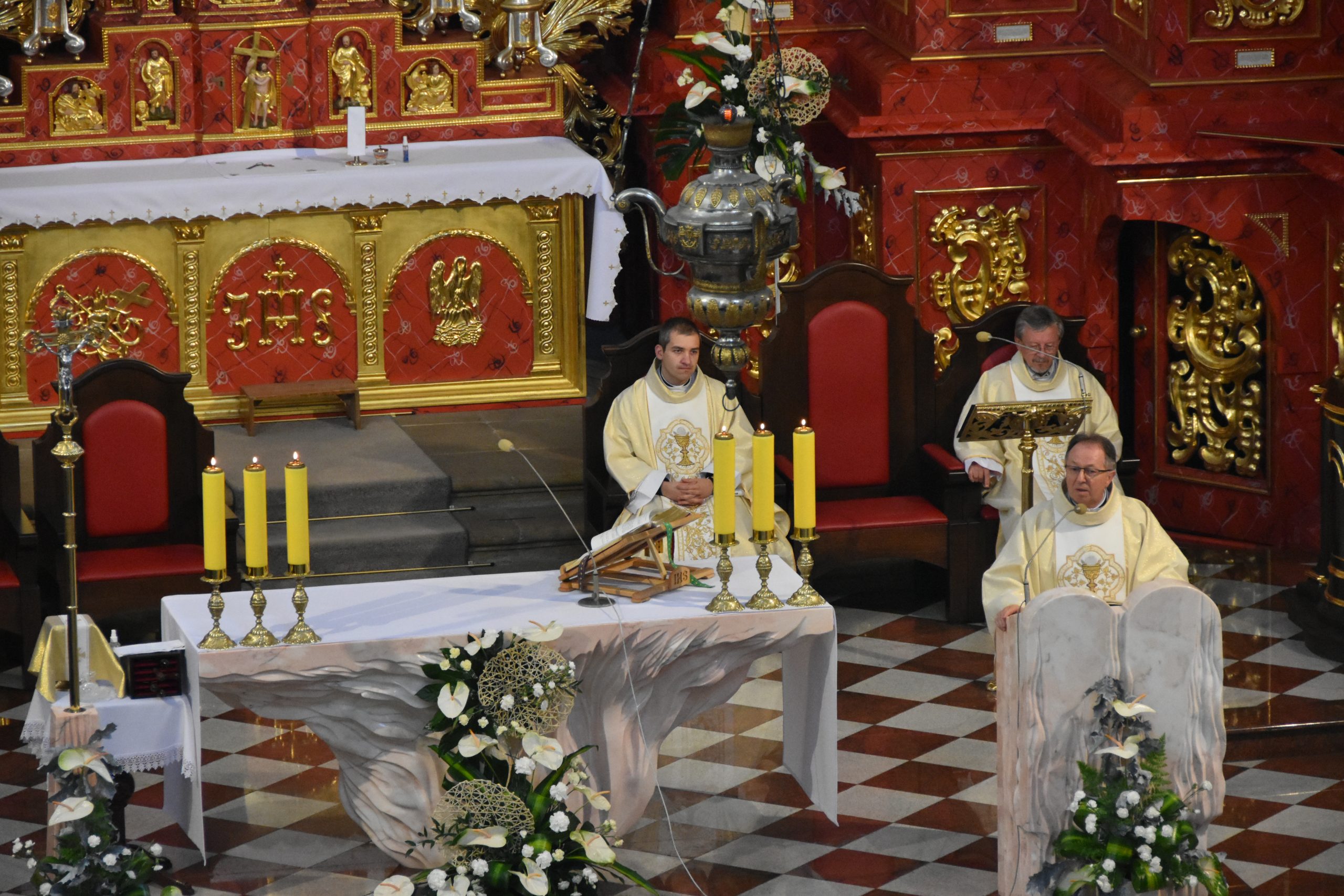 Stojący przy mównicy ksiądz ubrany w białą szatę ze złotymi ornamentami odprawia mszę świętą w intencji długoletnich małżeństw