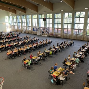 Grupa dzieci siedzą przy stolikach na sali gimnastycznej i grają w szachy,.