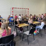 Grupa dzieci siedzi przy stolikach i gra w szachy na sali gimnastycznej.