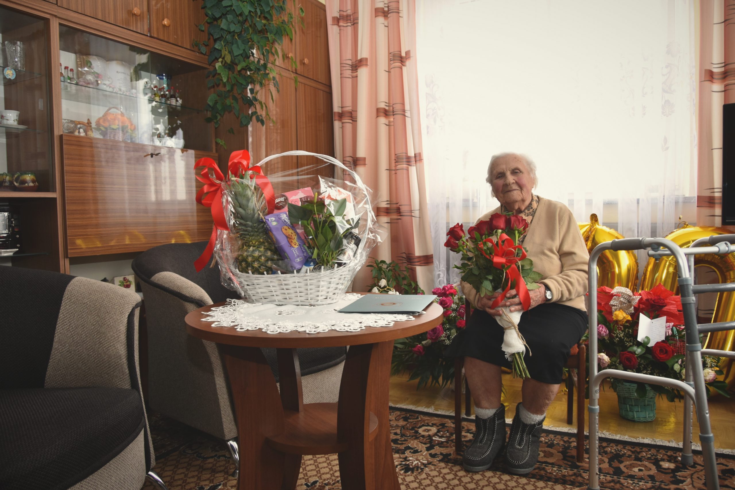 Uśmiechnięta, starsza pani z siwymi włosami ,znajdująca się po prawej stronie zdjęcia na tle zasłony, trzymająca czerwone róże. Na środku podarunki od urzędu miasta i gminy Brzozów.