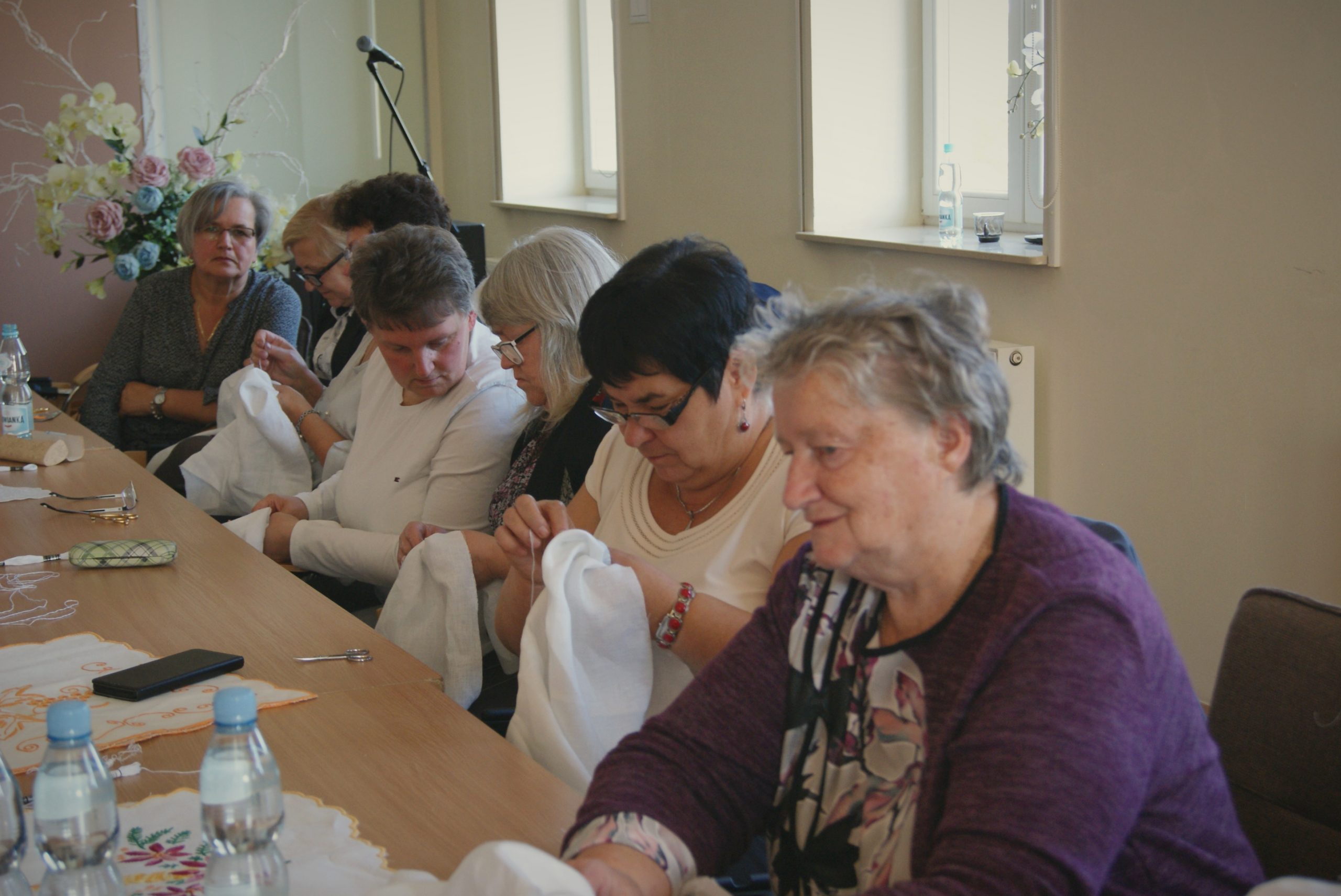 Grupa starszych kobiet siedząca przy stole i haftująca na dłutach.
