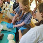 Grupa młodzieży podczas warsztatów kucharskich. Stoją w drewnianym pomieszczeniu, jedna z dziewcząt formuje ciasto.
