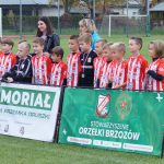Grupa młodych chłopaków ubranych w stroje piłkarskie stoi na murawie, za nimi dwie kobiety jedna młodsza o czarnych włosach, druga o blond włosach, przed nimi baner.