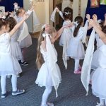 Grupa dziewczynek ubranych na biało tańczy w jasnej sali.