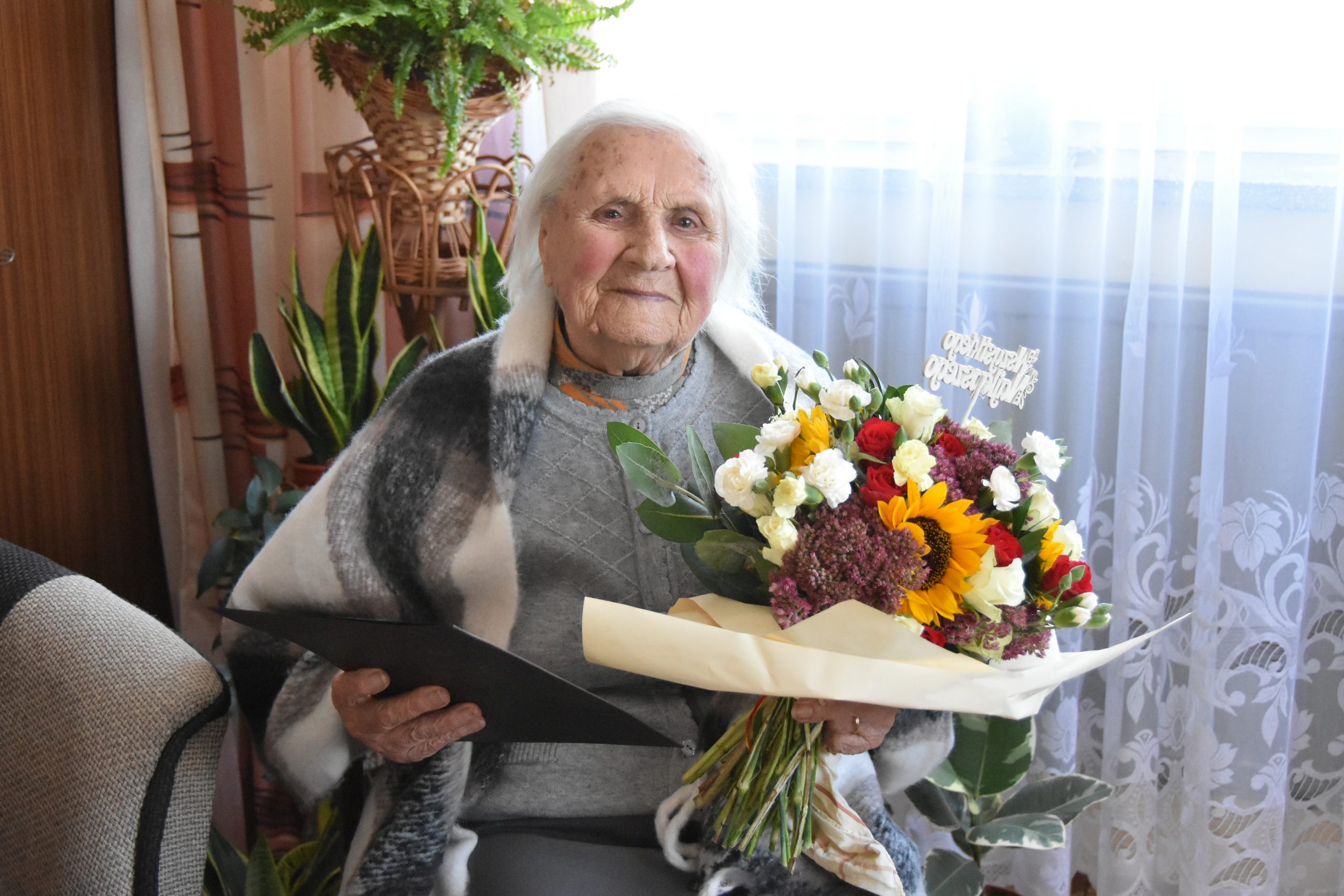 Starsza kobieta z siwymi włosami siedzi na krześle okryta szalikiem, trzyma w rękach bukiet kwiatów i teczkę z życzeniami.