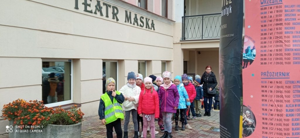 Grupa dzieci ubrana w kurtki i czapki stoi w parach przed jasnym budynkiem teatru.
