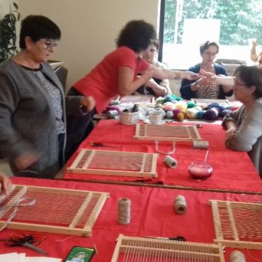 Grupa kobiet siedzi przy stole zasłanym czerownym obrusem i uczy się tkać na sprzęcie tkackim.