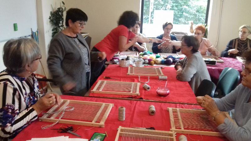 Grupa kobiet siedzi przy stole zasłanym czerownym obrusem i uczy się tkać na sprzęcie tkackim.