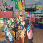Grupa dzieci tańczy w kolowerj sali pzredszkolnej, trzymając w rękach plasitkowe kółka.