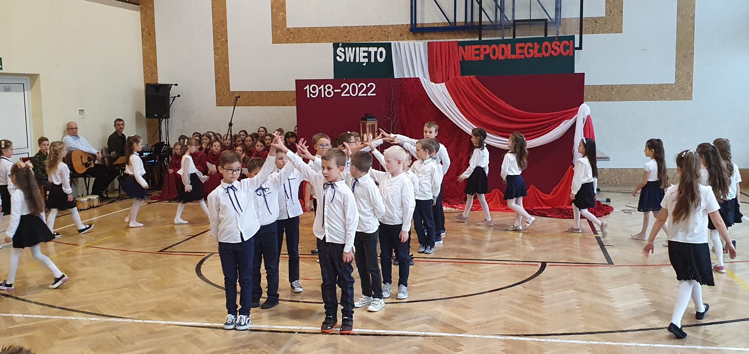 Grupa dzieci ubranych w odświętne stroje, białe koszule i ciemne spódnice, spodnie tancza poloneza na sali gimnastycznej.