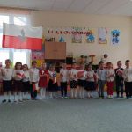 Grupa dzieci ubrana w odświętne stroje stoi na dywanie w sali szkolnej i śpiewa piosenkę. W rękach dzieci trzymają flagi biało czerwone. Za nimi na ścianie wisi duża flaga biało czerwona.