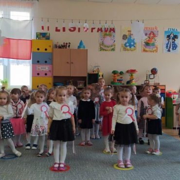 Grupa dzieci ubrana w odświętne stroje, z kotelionami biało czerownymi tańczy w sali szkolnej.