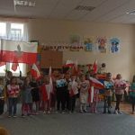 Grupa dzieci ubrana w odświętne stroje stoi na dywanie w sali szkolnej, w rękach dzieci trzymają flagę Polski, oraz prace konkursowe.