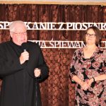 Starszy mężczyzna w ciemnym stroju księdza rzymskokatolickiego stojący wraz z kobietą w średnim wieku w kwieciestej sukience. W tle brązowa zasłona z białym napisem.