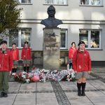 Grupa harcerzy stojąca przy popiersiu Józefa Piłsudskiego. W tle widać maszt i gmach rozległego budynku.