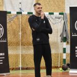 Młody wysoki mężczyzna ubrany w ciemny strój sportowy stoi na sali gimnastycznej i przemawia do mikrofonu.