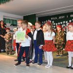 |Grupa dzieci ubranych w odświętne stroje mówi wierszyki na sali szkolnej. Na pierwszym pl;anie chłopiec trzymający w ręku mapę Polski.