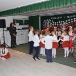 Grupa dzieci, dziewczynek i chłopaców ubranych w odświętne stroje tańczy w sali przedszkolnej.