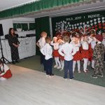 Grupa dzieci, dziewczynek i chłopaców ubranych w odświętne stroje tańczy w sali przedszkolnej.