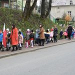 Grupa kilkudziesięciu dzieci wraz z kobietami idzie po chodniku. Na przodzie dwóch chłopaców trzyma w rękach flagi biało czerwone.