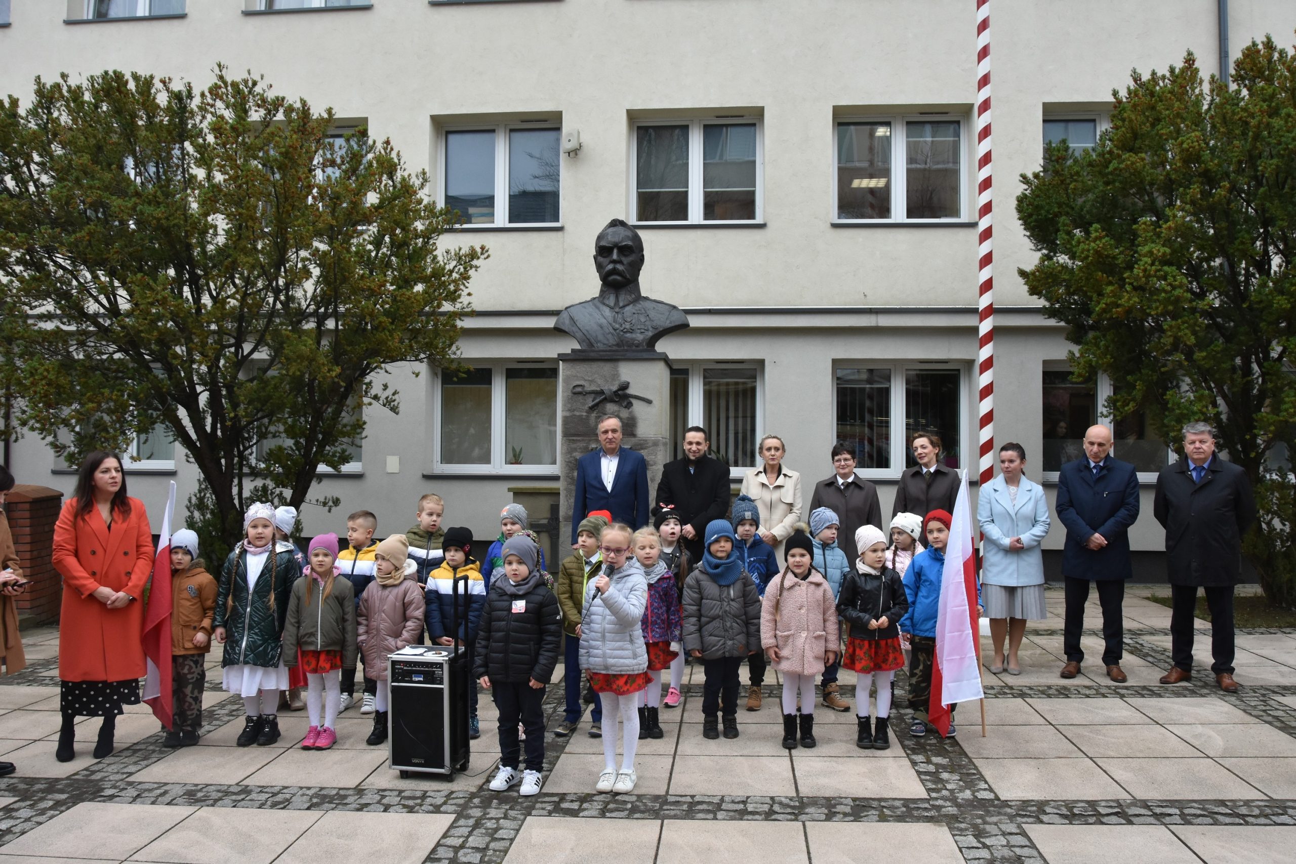 Grupa kilkudziesięciu osób (dzieci oraz kobiet i mężczyzn) stoją na kostce brukowej szarej przed jasnym budynkiem. Za nimi pomnik przedstawiające półpiersie Józefa Piłsdudskiego.