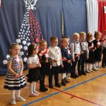 Grpa dzieci przedszkolnych obojga płci, stojąca przed ozdobną zasłoną z motywami patriotycznymi. W rękach trzymają bialo czerwone ozdoby. Znajdują się na rozleglej sali gimnastycznej.