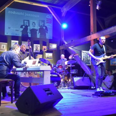 Grupa muzyczna złożona z czterech mężczyzn grajaca na instrumentach muzyczny na drewnianej scenie w rozległym pomieszczeniu.