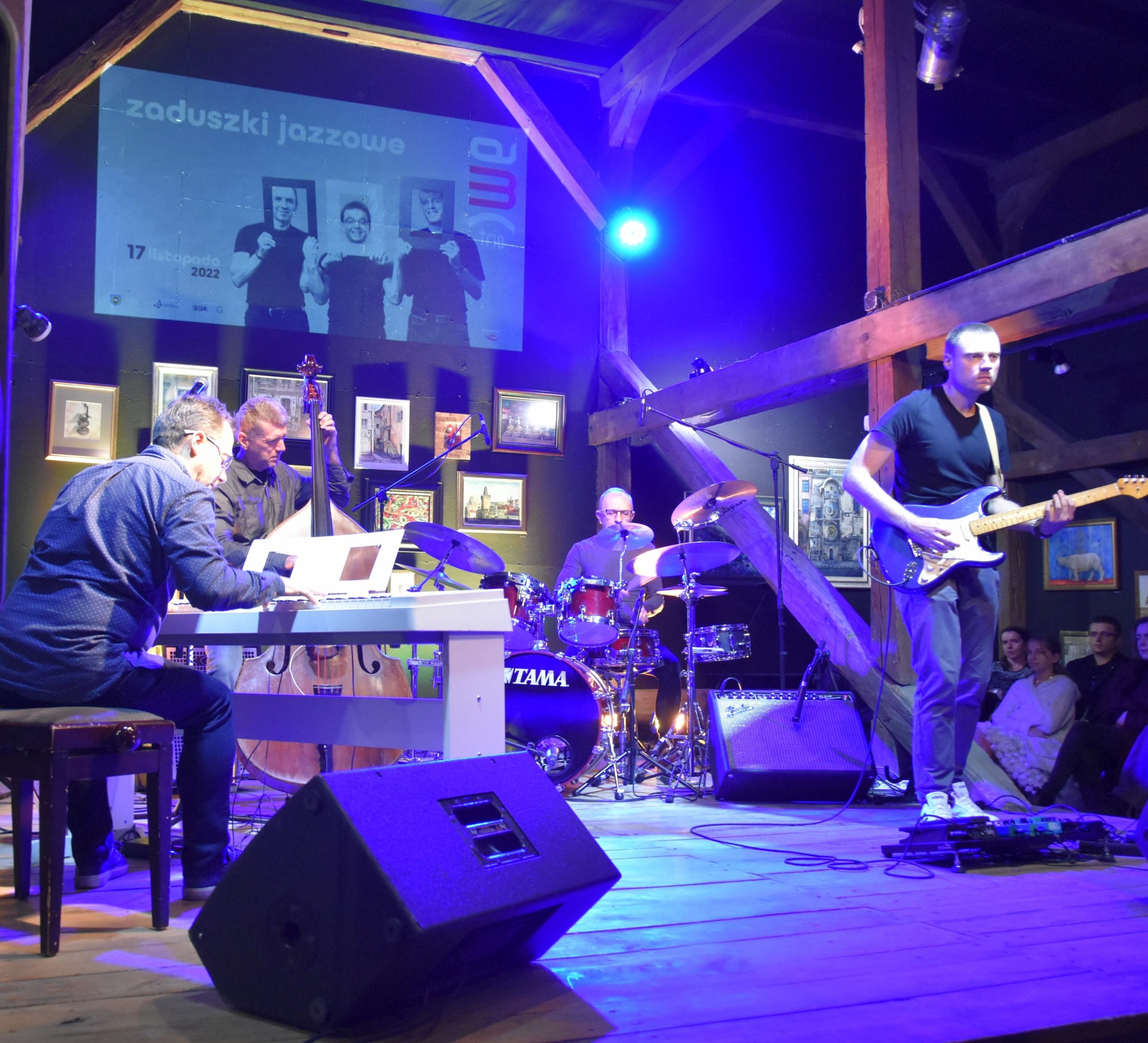 Grupa muzyczna złożona z czterech mężczyzn grajaca na instrumentach muzyczny na drewnianej scenie w rozległym pomieszczeniu.