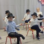 Grupa kilku chłopców w koszulach z kapeluszami na głowie, siedzi na krzesełkach w sali szkolnej i rusza rękami w rytm muzyki.