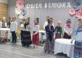 Grupa dzieci dziewczynek i chłopców przebranych w stroje długie spódnice i chusty stoi w sali przedszkolnej i recytuje wiersze. za nimi szara kurtyna z napisem Dzień Seniora.