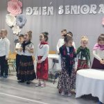 Grupa dzieci dziewczynek i chłopców przebranych w stroje długie spódnice i chusty stoi w sali przedszkolnej i recytuje wiersze. za nimi szara kurtyna z napisem Dzień Seniora.