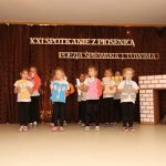 Grupa dzieci tańcząca na scenie przed ciemną kotarą.