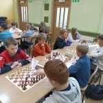 Grupa dzieci różnej płci i wieku grajaca w szachy na rozleglej sali.