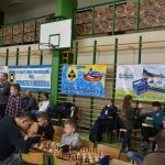 Grupa osób różnej płci i wieku na rozległej sali w trakcie rozgrywek szachowych. Na ścianie widać cztery banery.