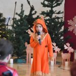 Dziewczynka w pomarańczowym stroju śpiewająca przed dorosłymi na sali gimnastycznej.