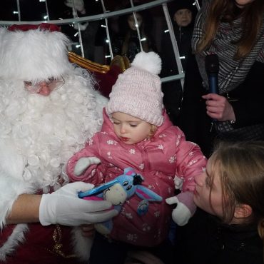 Młoda dziewczynka w zimowym stroju otrzymuje prezenty od św. Mikołaja. Wokół widać dwójkę dzieci.