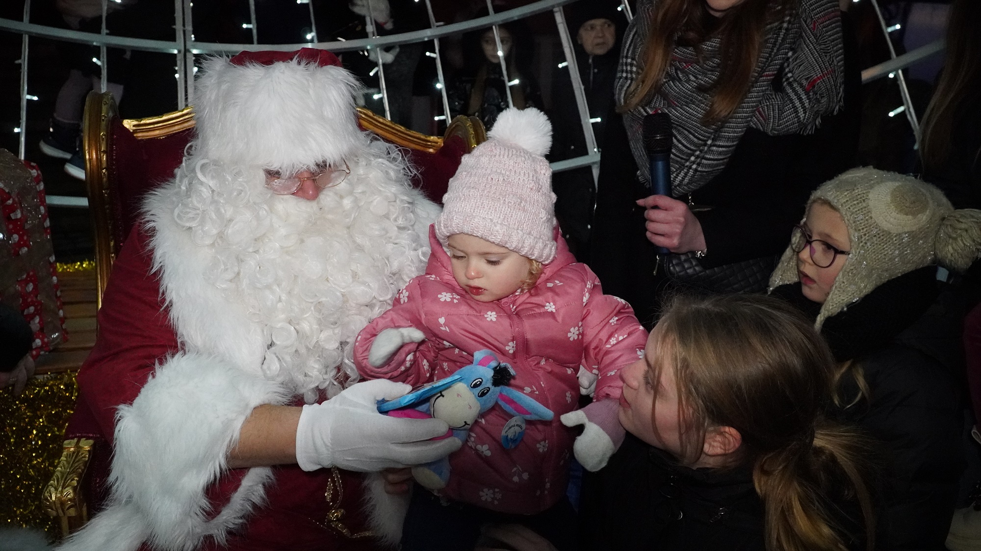 Młoda dziewczynka w zimowym stroju otrzymuje prezenty od św. Mikołaja. Wokół widać dwójkę dzieci.