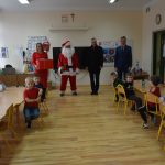 Mikołaj wraz z dwona dorosłymi mężczyznami i dorosłą kobieta trzymającą prezent w przedszkolu. z grupą dzieci.