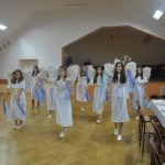 Grupa dziewczyn ubranych na biało ze skrzydełkami tańczą na sali z niebieskimi chustami.