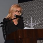 Kobieta w średnim wieku, włosy blond, ubrana w czarną marynarkę przewmawia do mikrofonu przy mównicy.