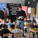 Grupa mężczyzn wśród dzieci grających w szachy na sali szkolnej.