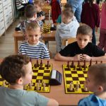 Grupa chłopców grających w szachy na sali szkolnej. W tle grupa dzieci.