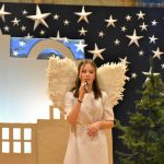 Dziewczynka w stroju aniołka śpiewa do mikrofonu. W tle ozdoby bożonarodzeniowe.