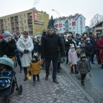 Grupa osób dorosłych i dzieci ubranych w zimowe kurtki idzie w przemarszu po ulicy.