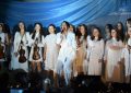 Grupa młodych dziewczyn ubranych na biało, stoją na scenie, za nimi niebieskie tło. Przed nimi stoi kobieta ubrana w biały kombinezon i spiewa do mikrofonu.
