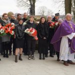 Grupa ludzi z bukietami kwiatów. Obok stoi dwóch duchownych w fioletowych ornatach.