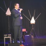 Mężczyzna w graniturze przemawia za pomocą mikrofonu na scenie.