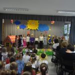 Grupa dzieci i dorosłych obserwująca występ teatralny przedszkolaków na salce szkolnej.