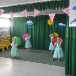 Grupa czterech dziewczynek z parasolkami na scenie w czasie wystepu artystycznego. Oglada je grupa przedszkolaków w żółtych stojach i nauczycielka.