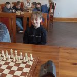 Chłopiec siedzący przy stoiku z szachami. W tle grupa dzieci grająca w szachy.